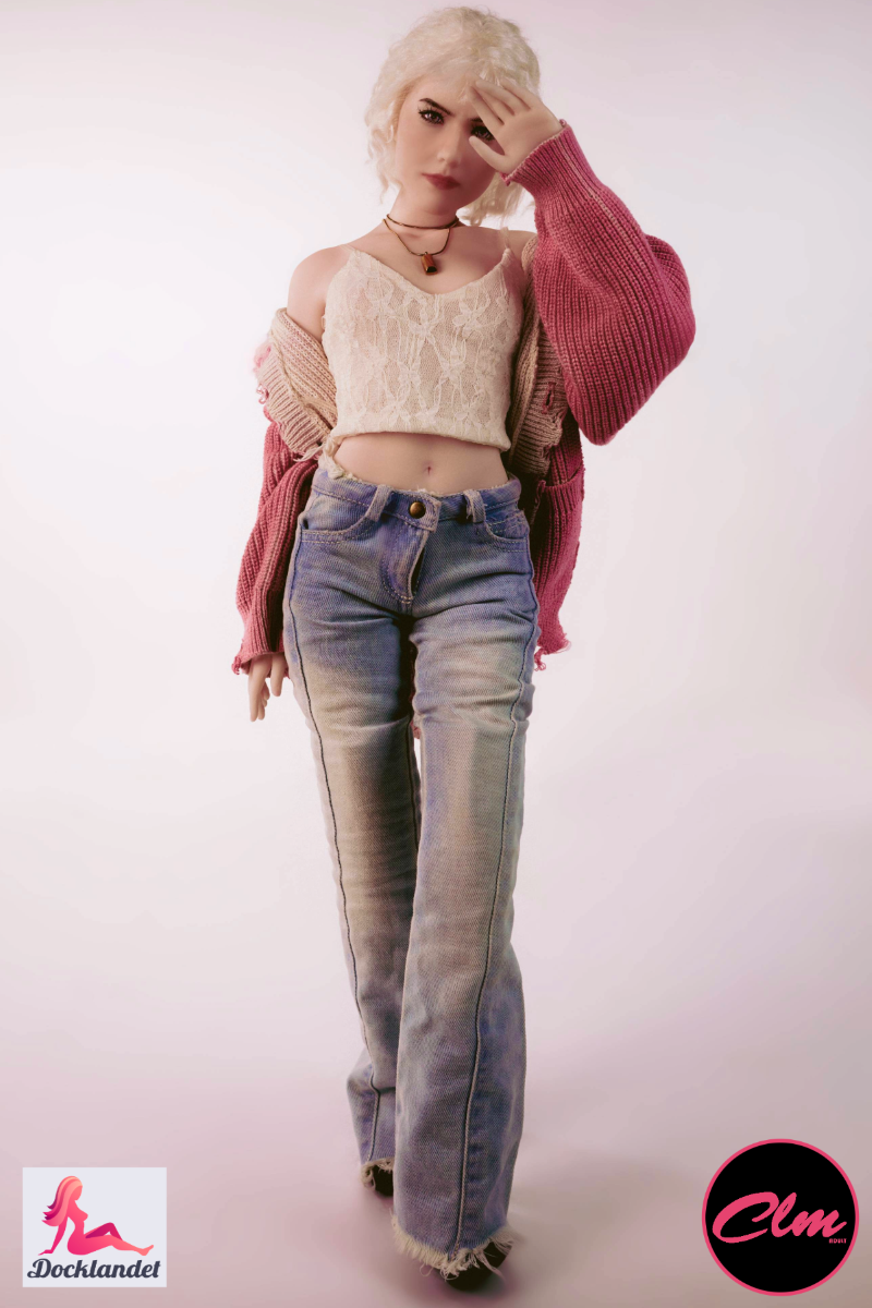 Sex doll Tara realistic mini 60 cm in silicone, blonde doll in jeans, crop top and pink jacket, standing upright against light studio background.